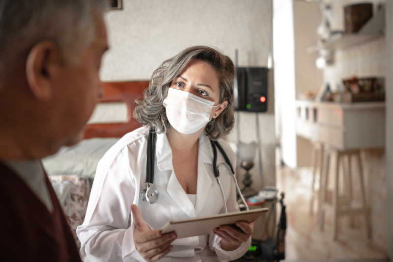 Masked female physician assistant wearing a white lab coat and stethoscope consults with an elderly male patient while holding a tablet.
