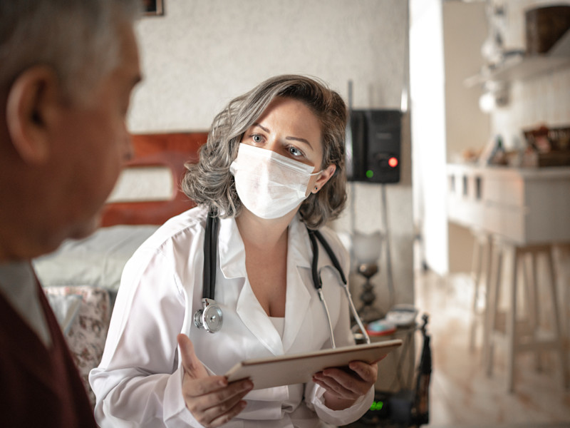 Masked female physician assistant wearing a white lab coat and stethoscope consults with an elderly male patient while holding a tablet.