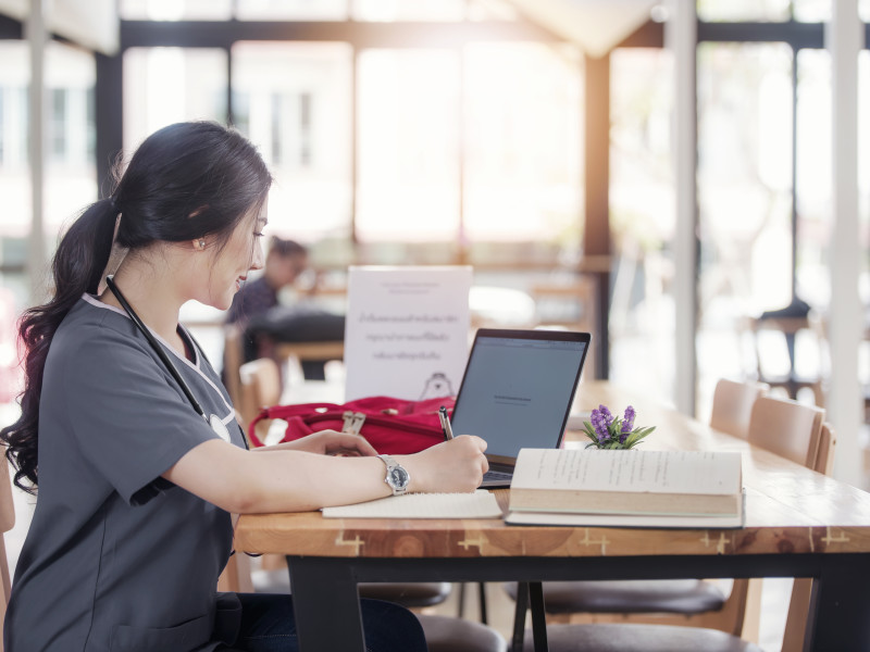 Female Asian-American nursing student wearing gray scrubs sits in a cafe doing her coursework on her laptop surrounded by books and notes.