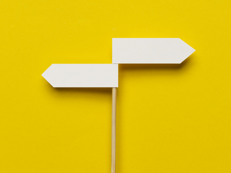 Skinny wooden pole with two white direction signs pointing in opposite directions against bright yellow backdrop.