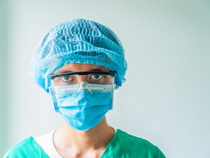 Female CRNA wearing a blue mask, blue hair covering, plastic goggles, and green scrubs stares directly into camera.