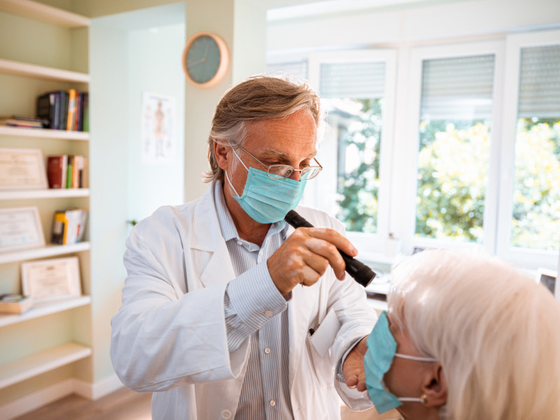 Older masked male optometrist with white hair shines light into elderly female patient while standing in a well-lit office.