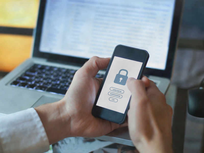 Hand holding a smartphone with a screen showing a locked icon with a laptop in the background.