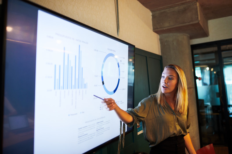 Young blonde businesswoman presenting financial results via a TV screen during a meeting.