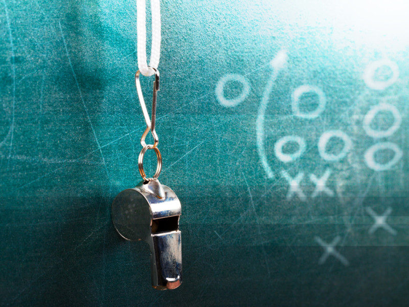 Silver coach whistle on a white string in front of blackboard with x's and o's drawn on it.