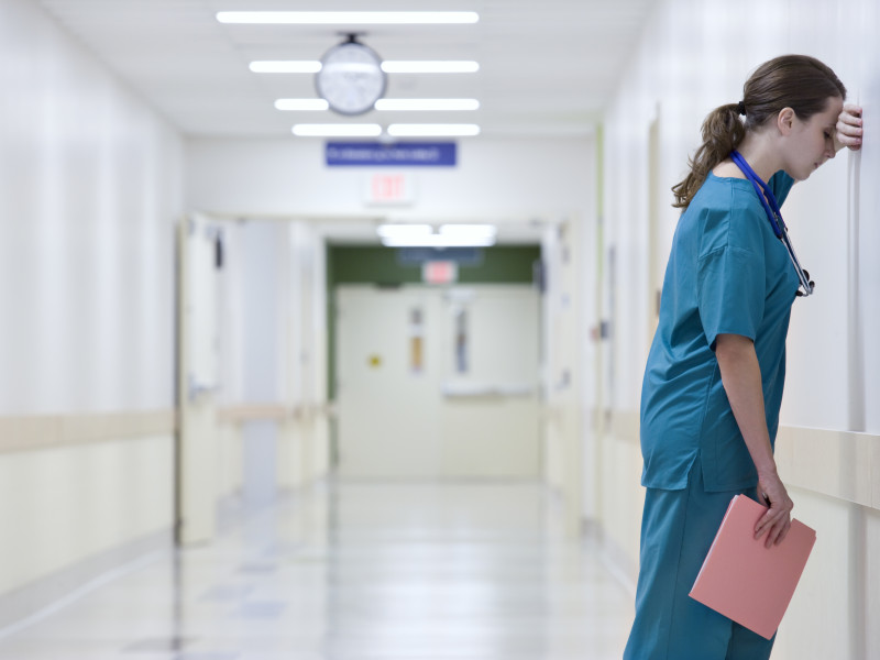 Stressed CRNA in blue scrubs holding a pink folder and leaning head against wall of white hospital hallway.