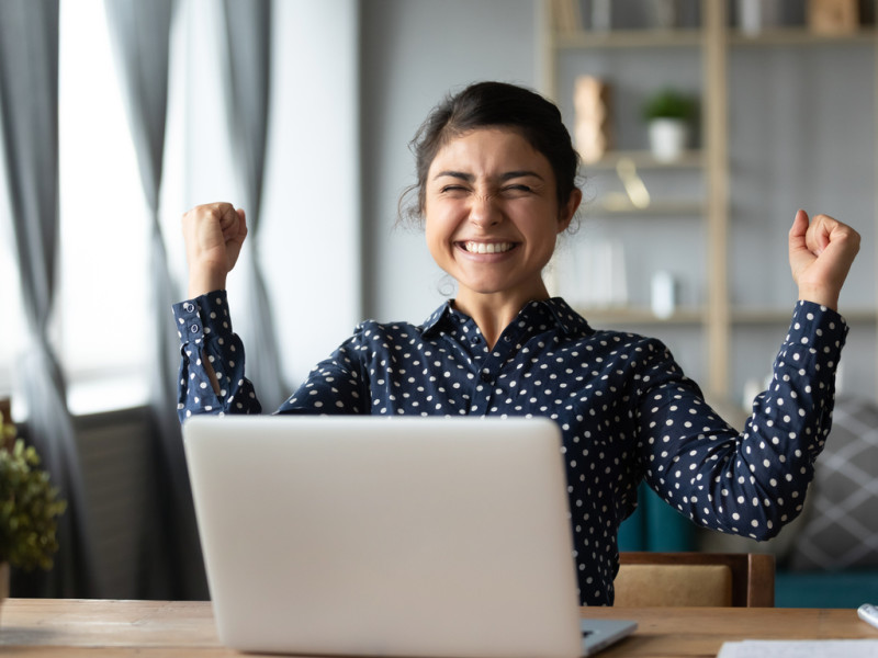 Young woman of color raising her arms in excitement while sitting in front of laptop.