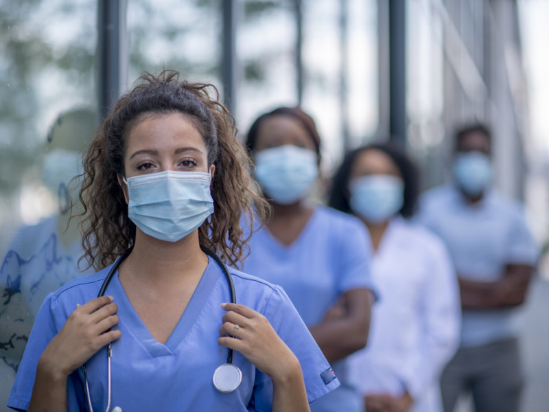 Masked female nurse practitioner standing outside facing camera with row of NPs standing behind her.