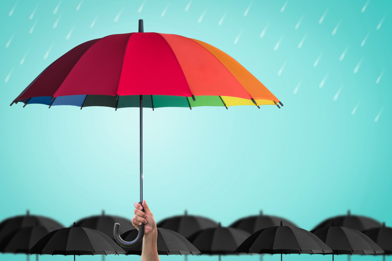 Hand holding rainbow umbrella above a sea of black umbrellas in the rain.