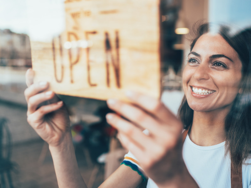 Young white woman with dark hair putting up a We're Open sign in her shop's front door