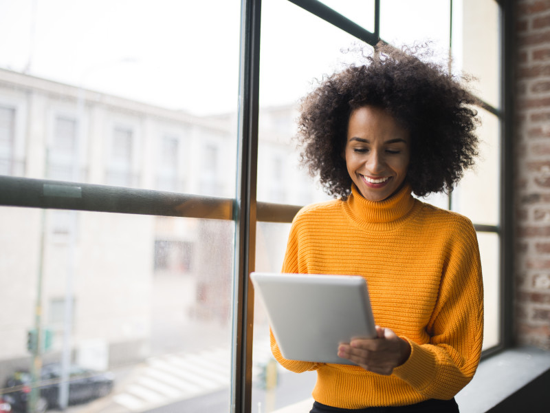一名非洲裔美国女房地产经纪人穿着橙色毛衣，微笑着浏览她的平板电脑。