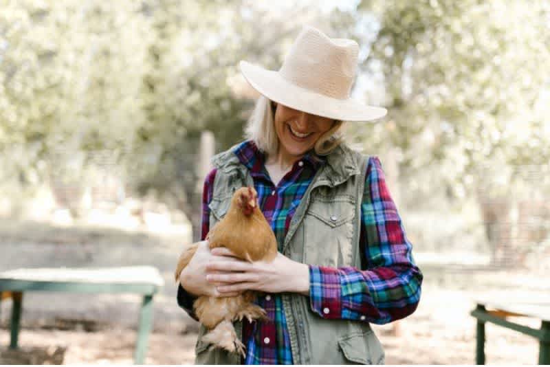 A woman wearing a hat with a plaid shirt and a vest outside holding a brown chicken and smiling down at it with trees in the background.