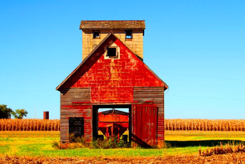 the front view of a red corn crib barn on a sunny day with corn fields in the background