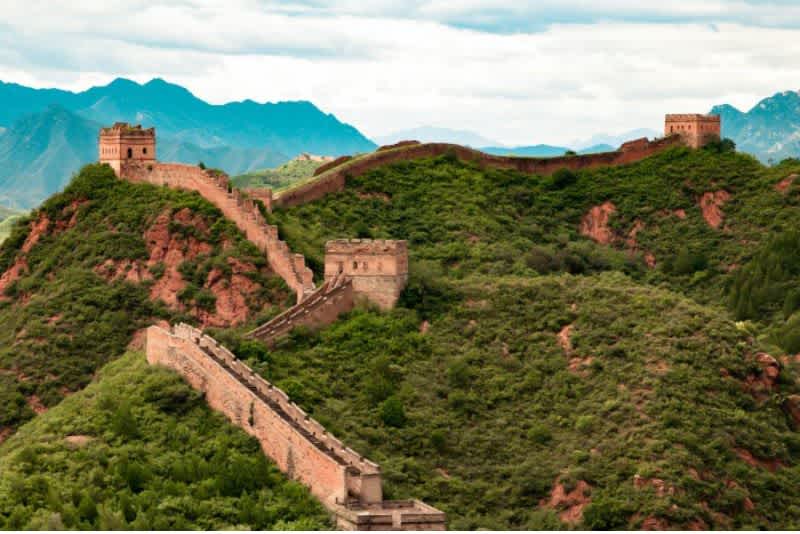 The Great Wall of China winds through green rocky hills with mountains and clouds in the background.