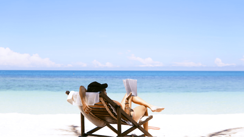 A woman wearing a hat reading a book while sitting on the beach