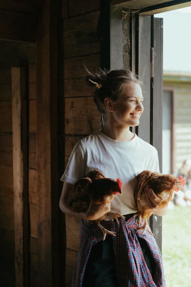 a smiling woman looking out the window holding two brown chickens