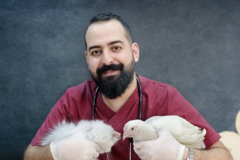 A veterinarian wearing red at a wooden table with a stethoscope around his neck, holding a white cat and a white chicken smiling.