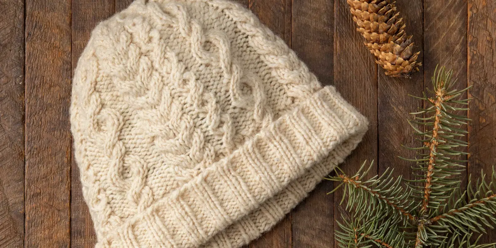 A cream-colored, handknit cabled hat rests on a wood surface next to an evergreen bough and pine cone.