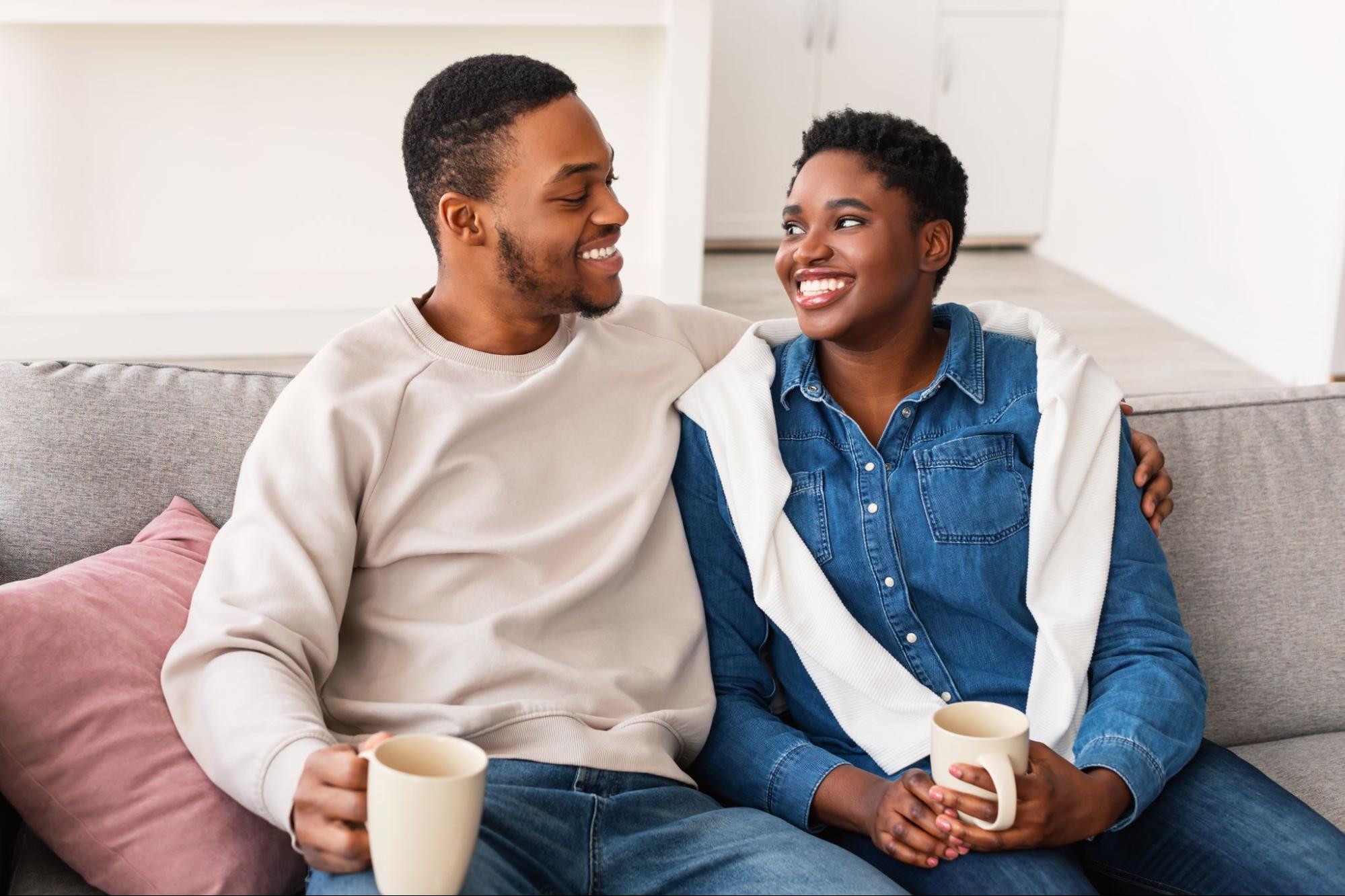 A smiling couple sits on their couch and drinks coffee