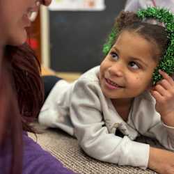 A student reads a book with a teacher while wearing a face shield