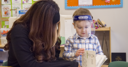 A student reads a book with a teacher while wearing a face shield