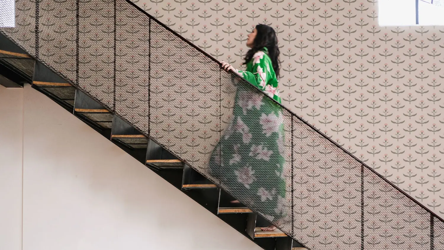 A woman in a green floral robe climbs a stairwell papered in Fine and Dandy wallpaper