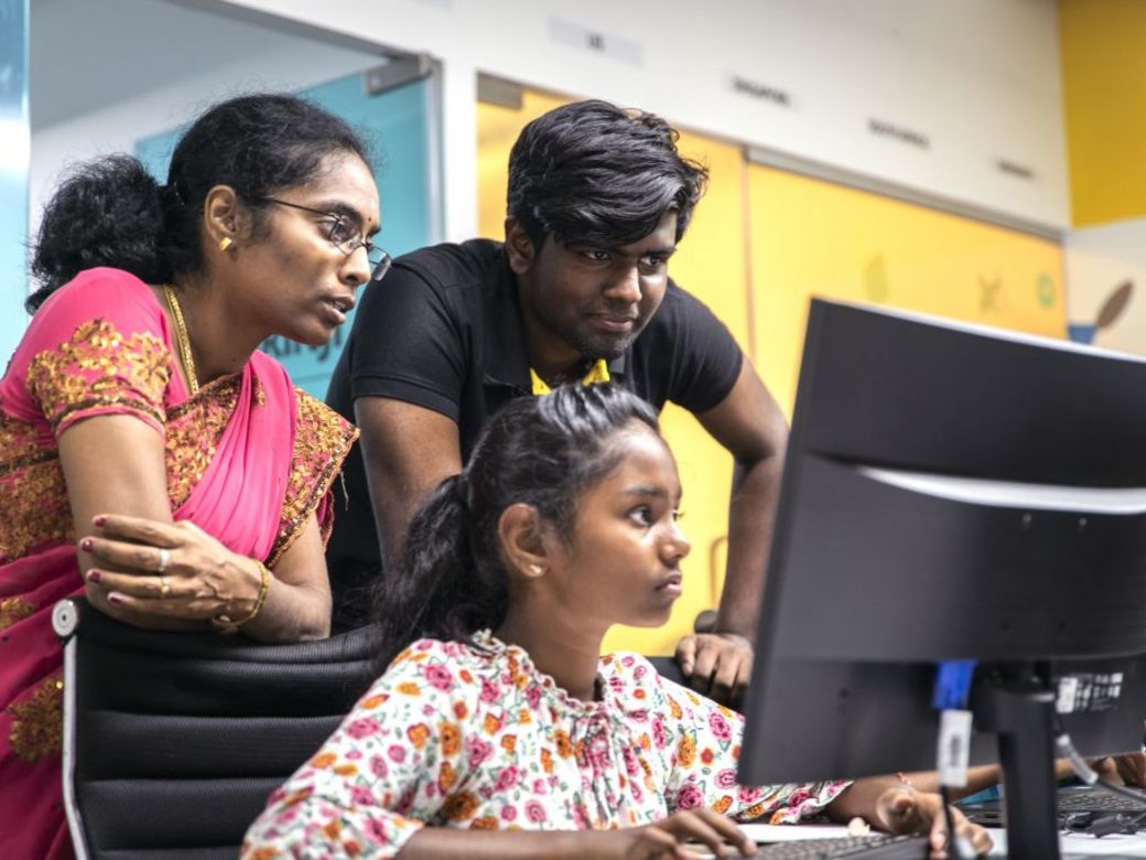 A girl sits at a computer with a male and female mentor standing behind her.