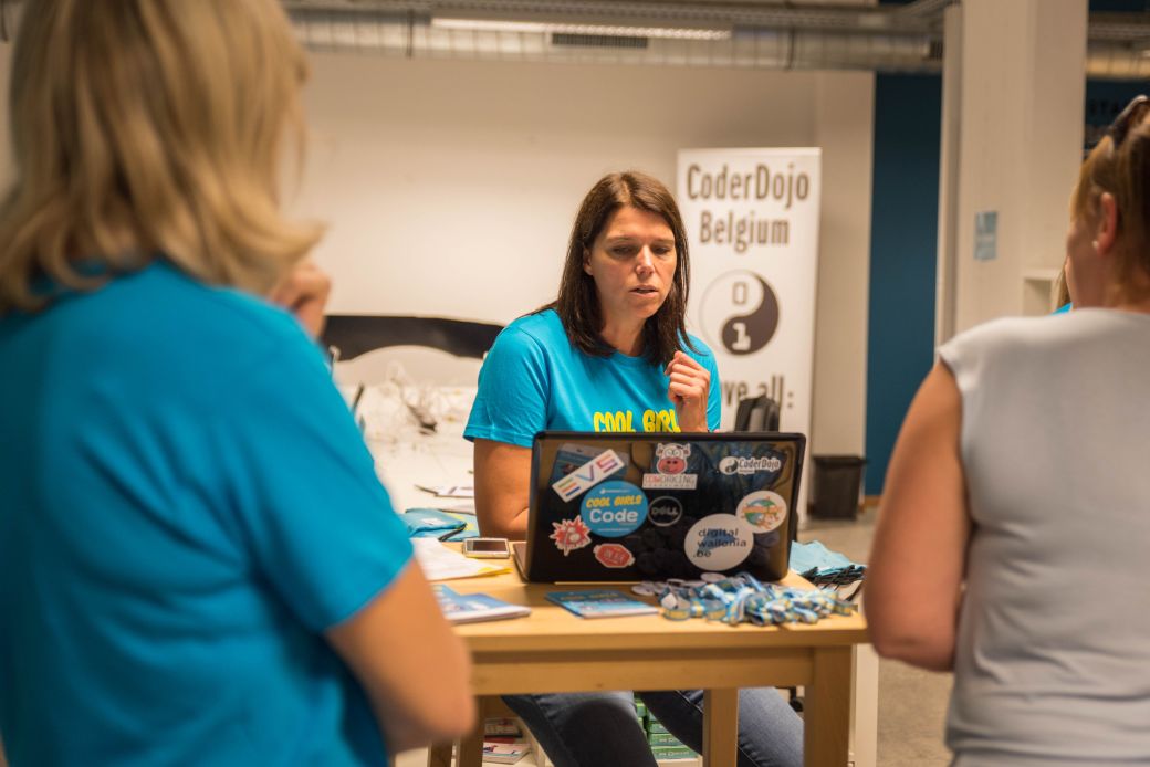 Group of three women standing around a table with a laptop on it. CoderDojo Belgium pull up banner in the background.