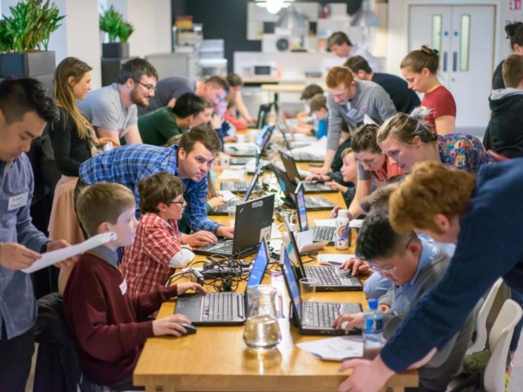 Mentors and young people on laptops coding on a long table. Image credit: Marie Greene