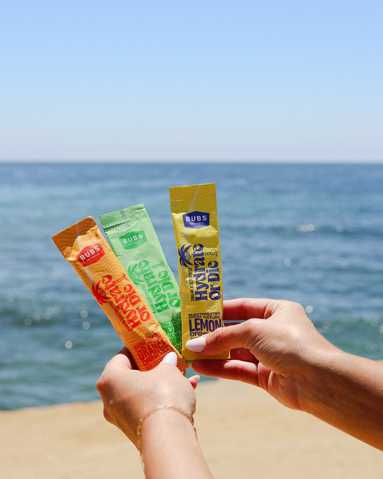 BUBS Naturals Hydrate or Die Electrolyte Packets being held above the ocean, orange coconut and lemon flavors