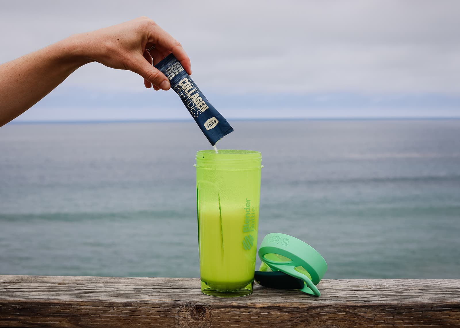 BUBS Naturals Collagen Peptides being poured into a green blender bottle overlooking the ocean