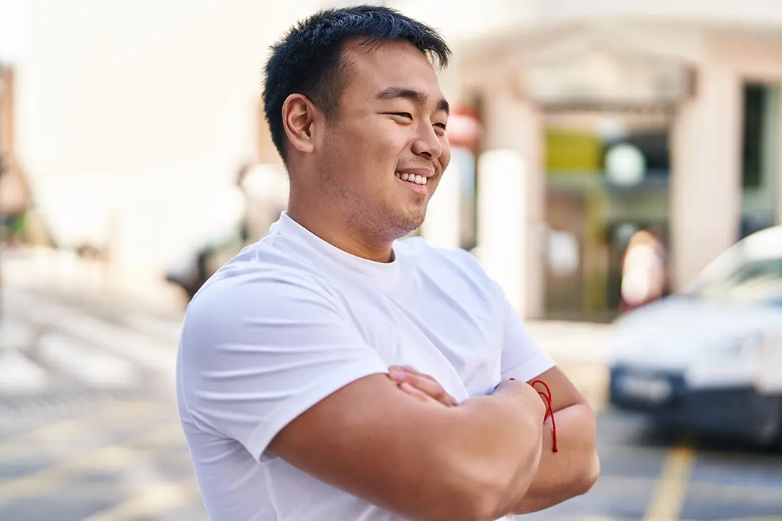 A young asian man standing on the street with his arms folded.
