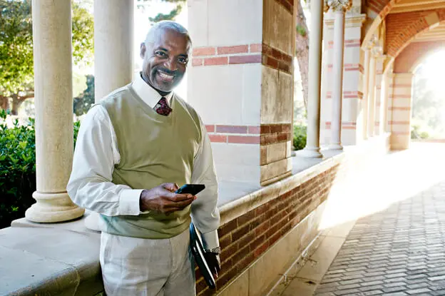An elderly afro-american man is standing inside a campus corridor holding his phone.