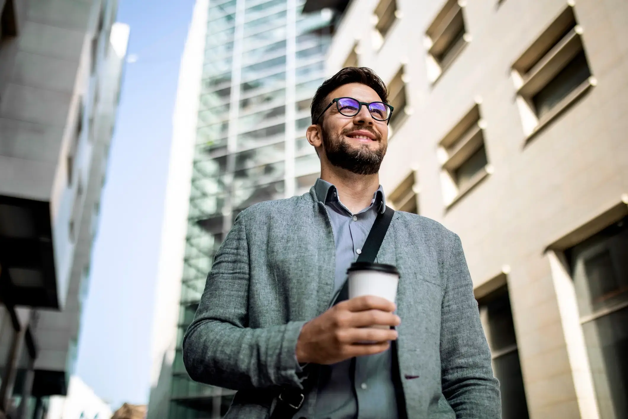 A business man walking outside amongst tall buildings with a coffee.