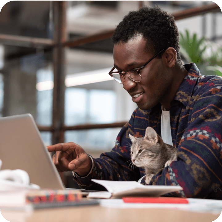 A man cradling his cat while pointing at a laptop screen, representing the use of supported browsers by Embrace Pet Insurance for seamless online access and pet insurance management.