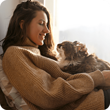 A smiling woman who is looking so relieved that she acquired pet insurance for her beautiful long-haired cat sitting on her lap and lovingly gazing at her. 