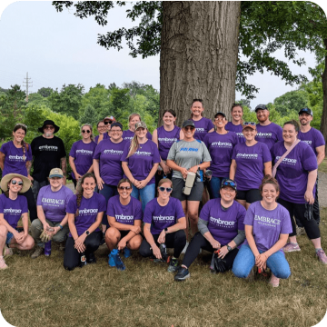 Embrace Pet Insurance team members in purple shirts gather outdoors under a large tree, showcasing their diverse and engaged workforce participating in a company event or volunteer activity, highlighting the collaborative and community-oriented culture.