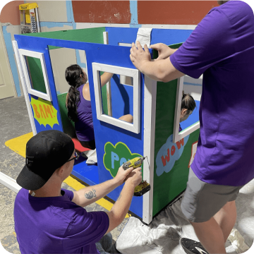 Embrace Pet Insurance employees in purple shirts collaboratively paint a colorful playhouse, demonstrating teamwork and community involvement, showcasing the company's commitment to charitable activities and fostering a positive work culture.