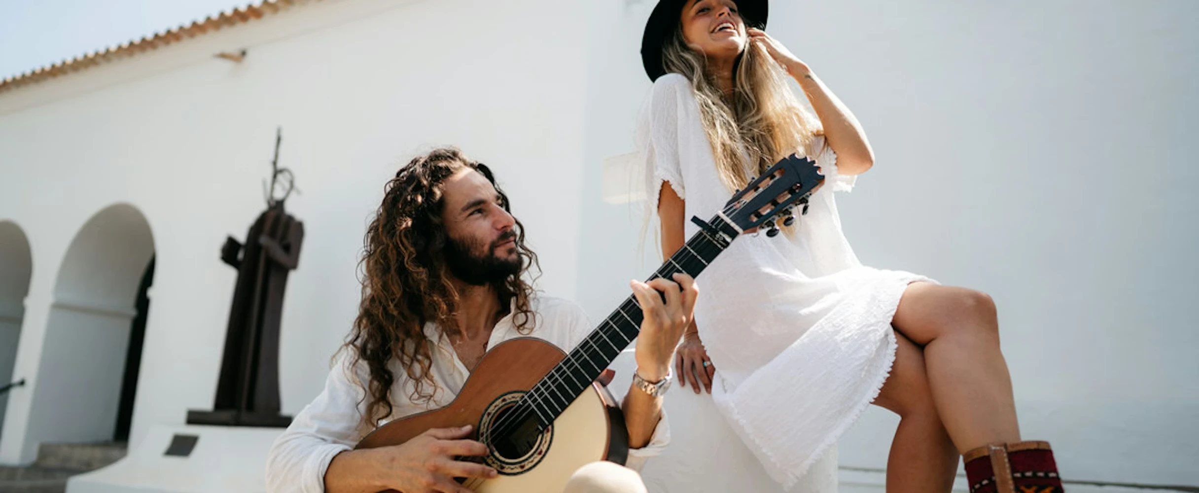 A man with long curly hair playing an acoustic guitar while looking up at a smiling woman in a white dress and black hat, seated outdoors against a bright white building.