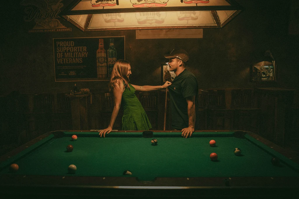 A man and woman stand beside a pool table, engaged in conversation, with cues and balls visible on the table.