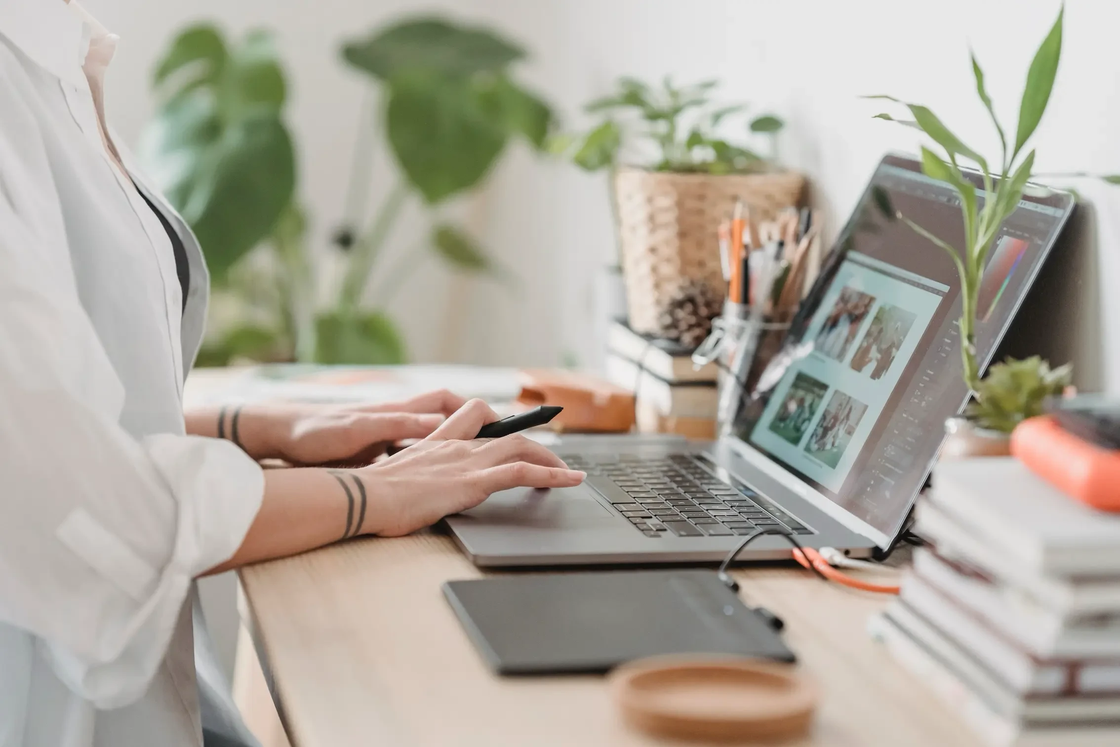 Close-up side view of a photographer's hands using a laptop and stylus to edit a gallery of images in a bright workspace.