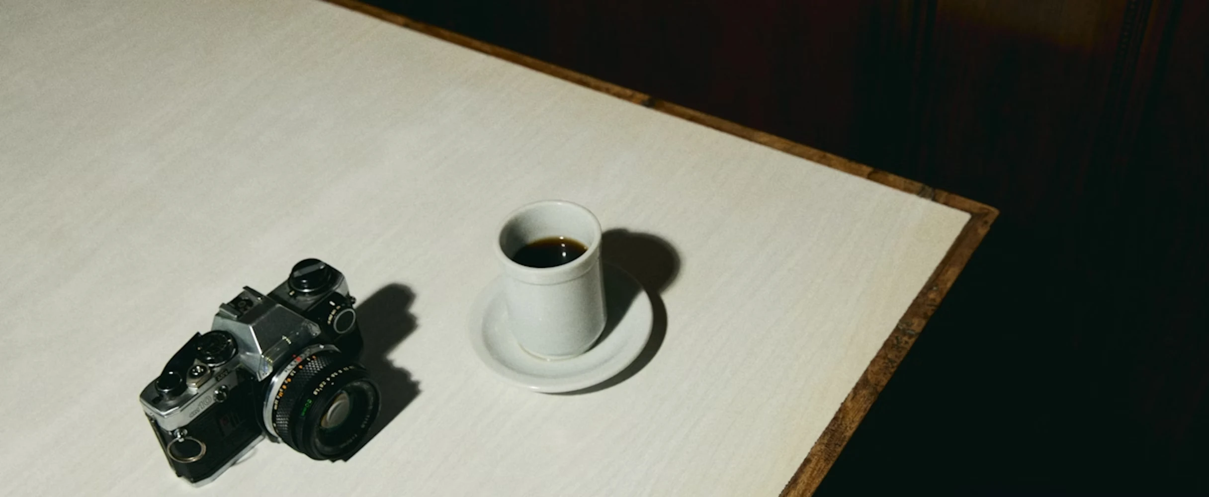 Overhead photo of a film camera and a small white cup of black coffee on a light-coloured table with dark background for a simple, moody workspace scene.