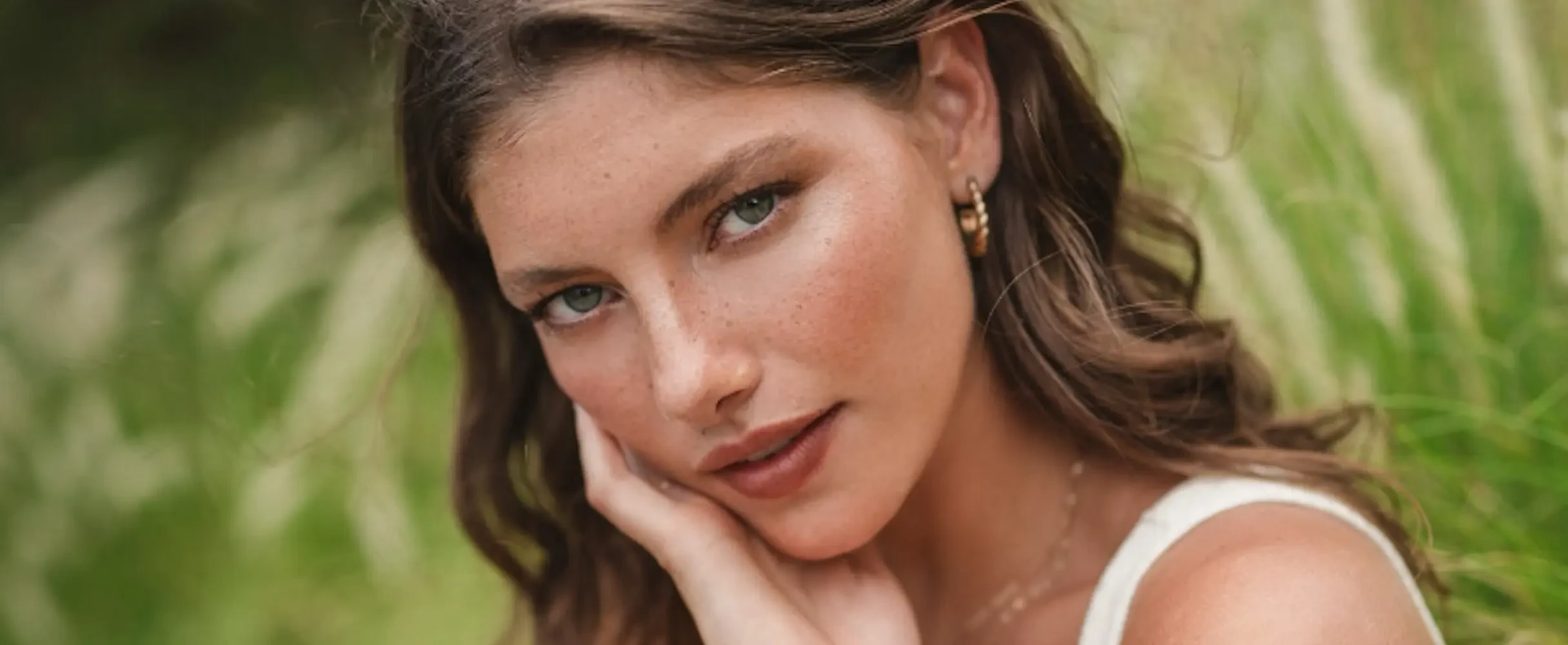 Close-up outdoor portrait of a young woman with freckles and wavy brown hair, resting her hand on her chin against a soft, blurred green grass background.