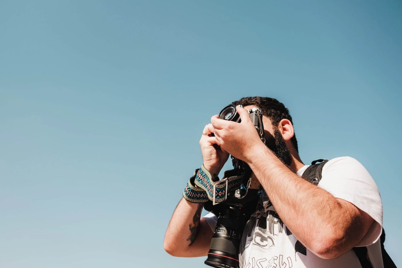 A man takes photos with a camera against a clear blue sky. Daytime outdoor photography scene. Photo by Airam Dato-on: https://www.pexels.com/photo/sunlit-man-taking-pictures-15189445/