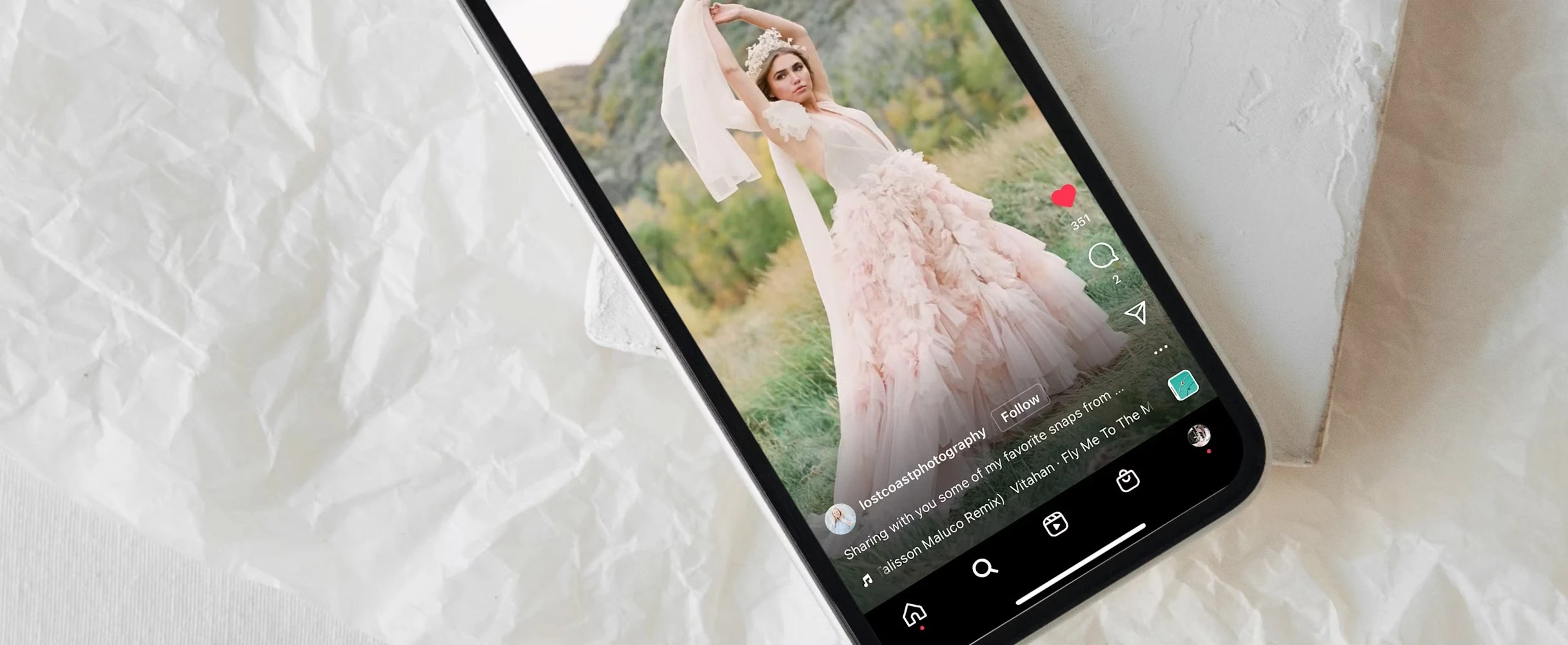 A smartphone rests on a textured white surface, showing a social-media post of a woman outdoors wearing a light pink, ruffled gown and a floral headpiece, posing with her arms raised against a natural landscape.
