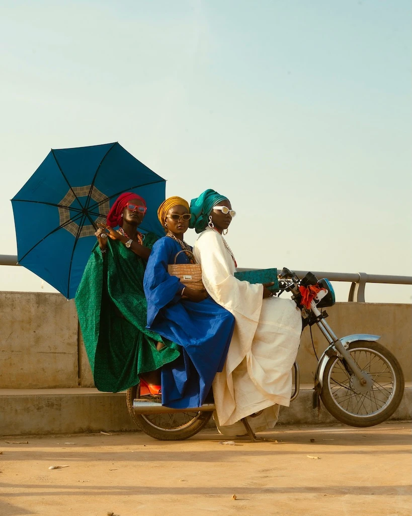 Three people with styled hair (red, blonde, teal) wearing vibrant colored garments (green, blue, white) riding a motorbike on an elevated concrete platform, sharing a blue umbrella. Photo By Etim Essang.