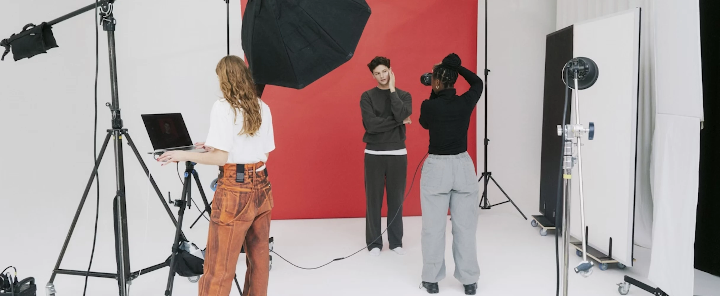 A photographer takes pictures of a person posing in front of a red backdrop, while another crew member stands nearby working on a laptop. Studio lighting equipment and reflectors surround the setup in a bright, minimalist photo studio.