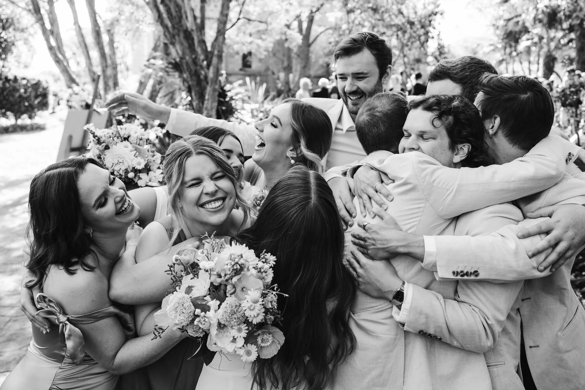 Black and white close-up shot of a joyous wedding party embracing in a tight group hug while laughing and holding flower bouquets. Image by Translucent Photography.
