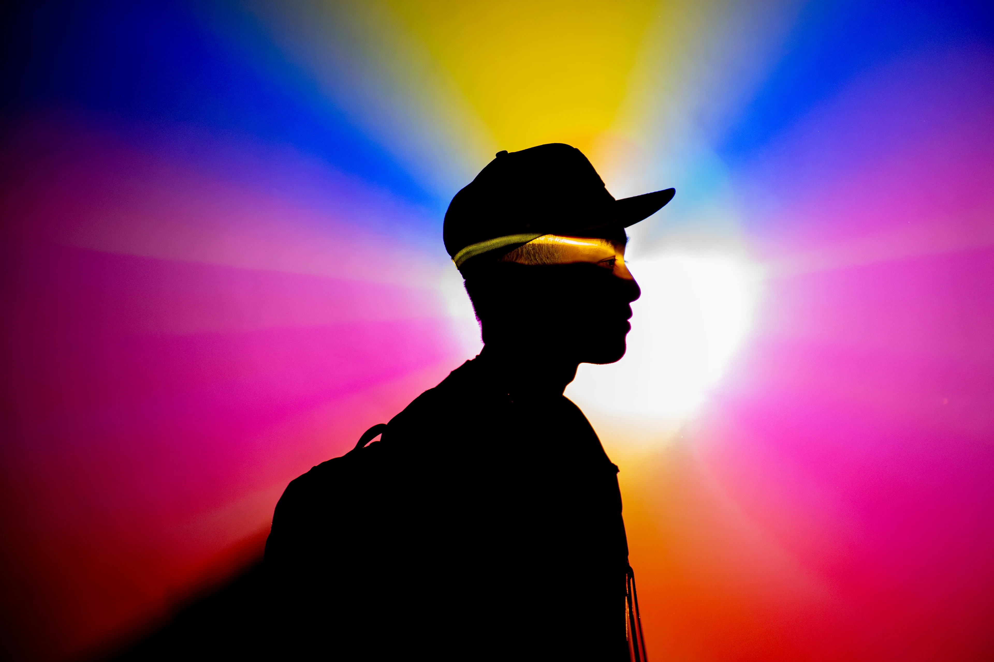 Chromatic Profiling. The profile of a silhouetted figure standing in front of a multi-colored glow. Photograph captured by Tyler McCormick at MODE Festival by Flickr (https://www.flickr.com/photos/157060264@N02/55539117255/)