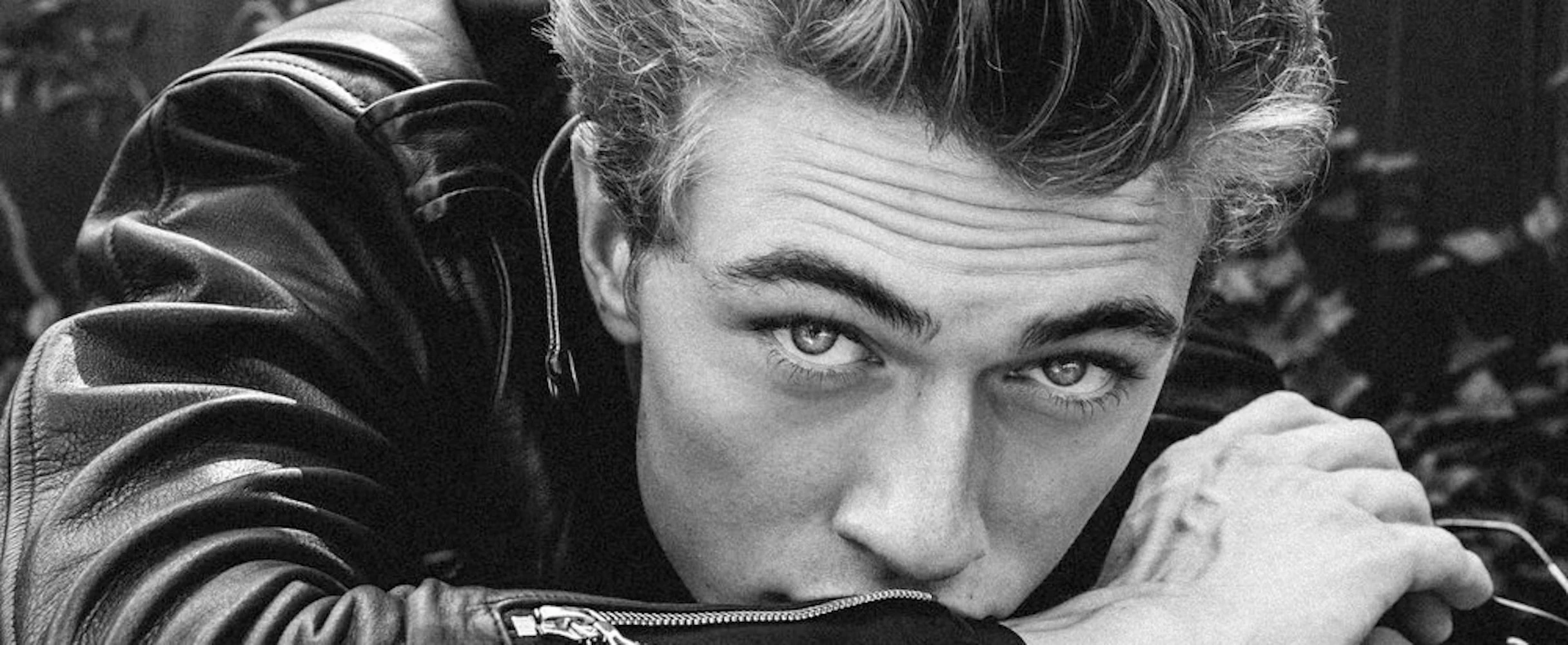 A close-up black and white photograph of a young man with styled hair wearing a leather jacket, resting his chin on his arm and looking directly at the camera. Photo By Katie Levine